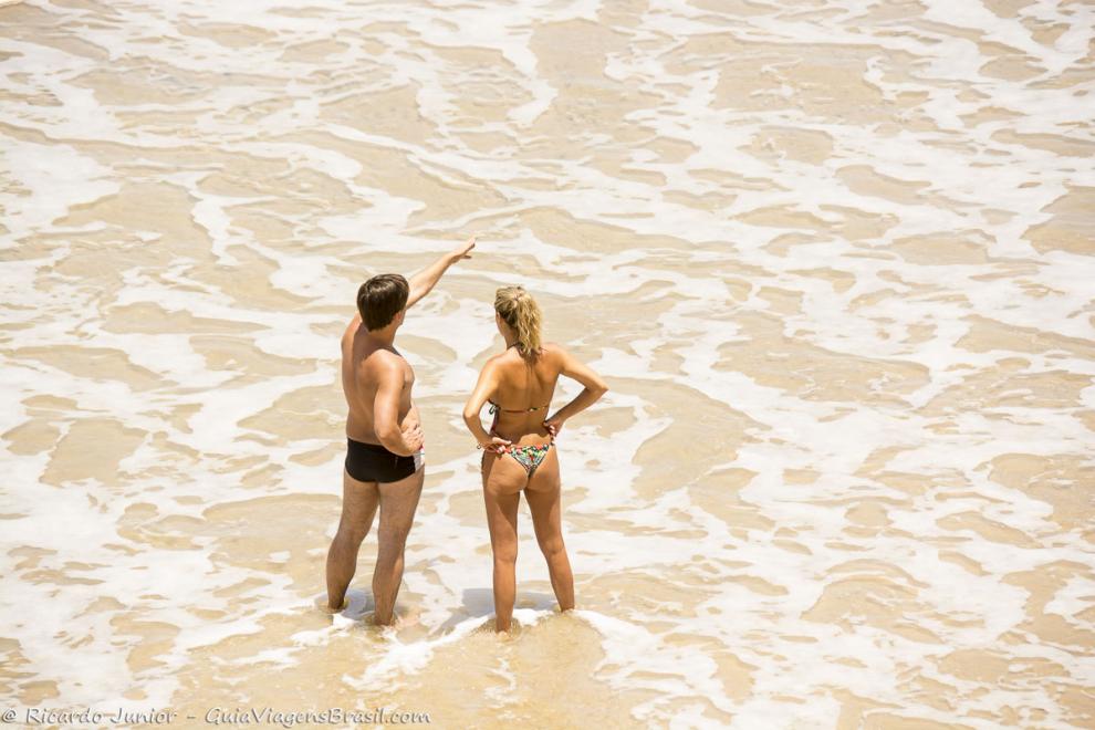 Imagem casal conversando com pés nas águas da Praia da Engenhoca.